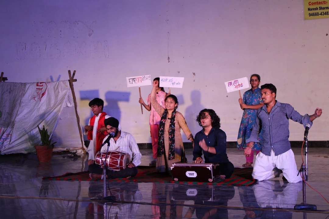 An ensemble performs with raised hands, placards, a drum and a harmonium